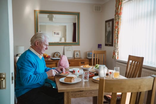 One senior man is sitting at the dining table in his home having lunch. He is making a ham salad sandwich.
