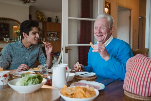 Senior man is having lunch at home with his grandson. They are eating sandwiches and potato chips.