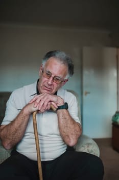 Senior man is looking sad with his head resting on his walking stick in the living room of his home.