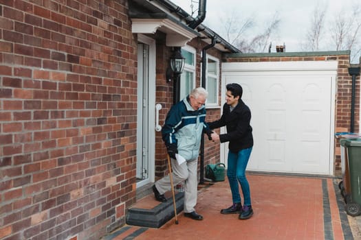 Teenage boy is helping his grandfather out of the door and down the steps of his house.