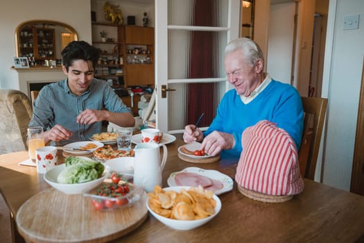 Senior man is having lunch at home with his grandson. They are eating sandwiches, pizza and potato chips.
