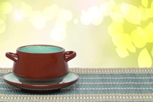Table top on bowl background. Empty brown ceramic soup bowl on plate on a green bamboo straw mat over abstract bright yellow spring backdrop. Template for your display product montage.