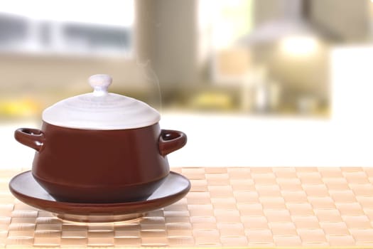 Table top on bowl background. Closeup of hot brown ceramic soup bowl on plate with lid on a bright tablecloth over blurred kitchen interior background. Template for your display product montage.