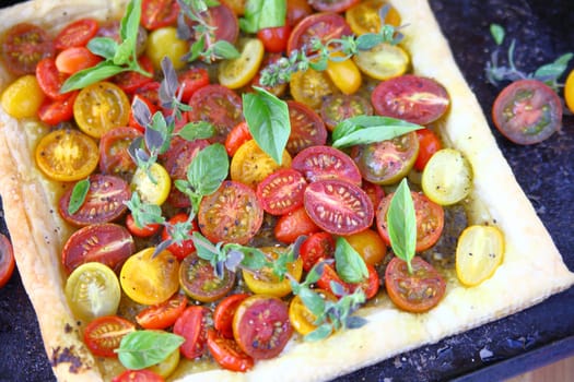 Overhead view of cherry tomato tart on puff pastry with basil and oregano leaves