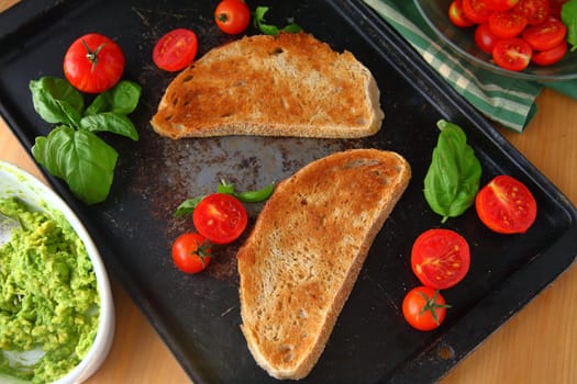 Overhead view of toast slices, mashed avocado, cherry tomatoes and basil on old baking pan
