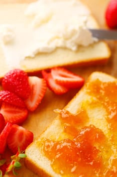 Spreading cream cheese on white bread with jam and strawberries alongside