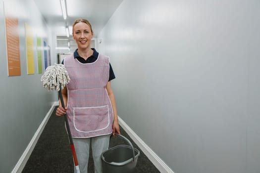 Portrait of a mature female cleaner with a mop and bucket in the corridor of an office building.