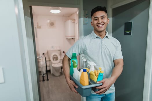 Portrait of a young, male cleaner at work. He is holding a tray of cleaning products.