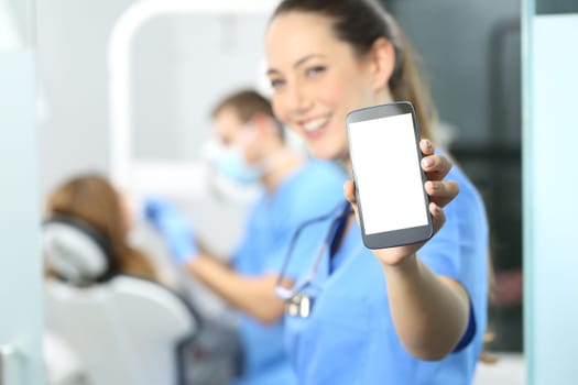 Stomatologist showing phone screen and looking at you in a dentist office interior with a doctor working in the background