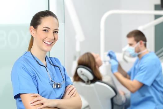 Dentist female with crossed arms wearing blue coat posing and looking at you at consultation with a doctor working with a patient in the background