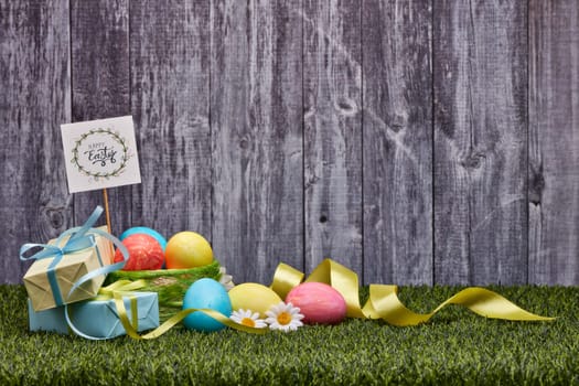 Colored easter eggs, gift boxes and flowers lying on a green grass on a grey wooden background with copy space. Greeting card, Easter background