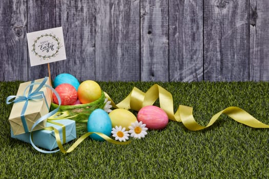 Colored easter eggs, gift boxes and flowers lying on a green grass on a grey wooden background with copy space. Greeting card, Easter background