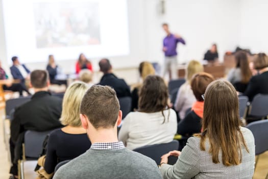 Business and entrepreneurship symposium. Speaker giving a talk at business meeting. Audience in the conference hall. Rear view of unrecognized participant in audience.