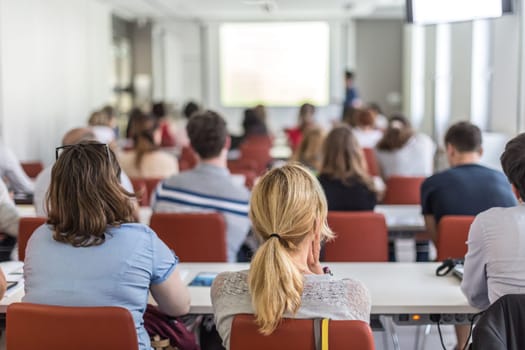 Female speaker giving presentation in lecture hall at university workshop. Audience in conference hall. Rear view of unrecognized participant in audience. Scientific conference event.