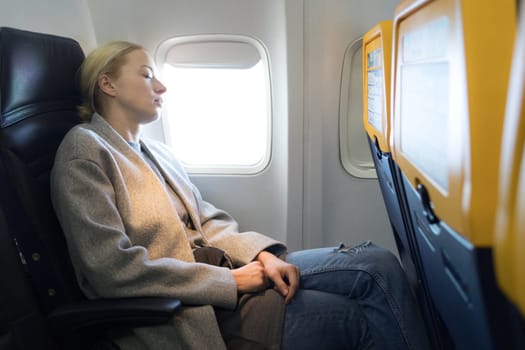 Tired blonde casual caucasian woman wearing glasses, napping on seat while traveling by airplane. Commercial transportation by planes.