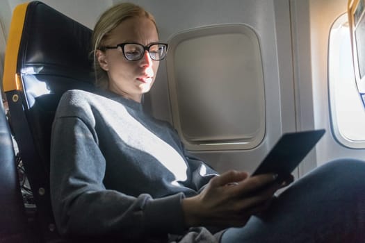 Thoughtful blonde casual caucasian woman wearing glasses reading on digital e-reader while traveling by airplane. Commercial transportation by planes.