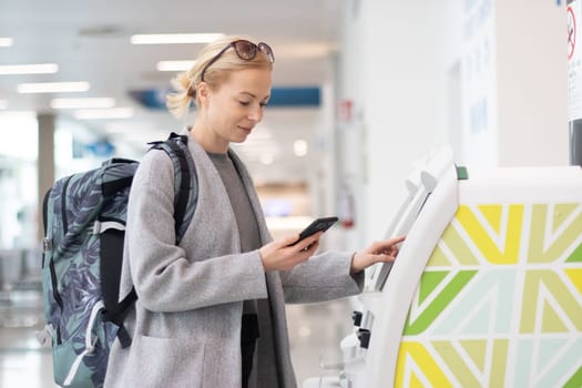 Casual caucasian woman using smart phone application and check-in machine at the airport getting the boarding pass. Modern technology on airport.