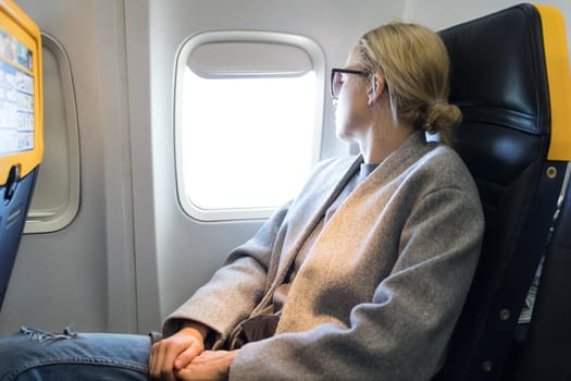 Thoughtful blonde casual caucasian woman wearing glasses, looking through the window while traveling by airplane. Commercial transportation by planes.