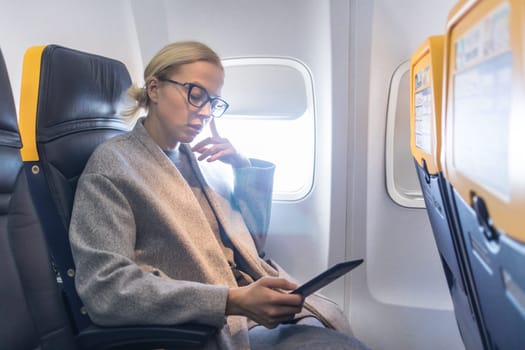 Blonde casual caucasian woman wearing glasses reading on digital e-reader while traveling by airplane. Commercial transportation by planes.