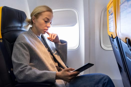 Thoughtful blonde casual caucasian woman reading e-book on digital e-reader while traveling by airplane. Commercial transportation by planes.