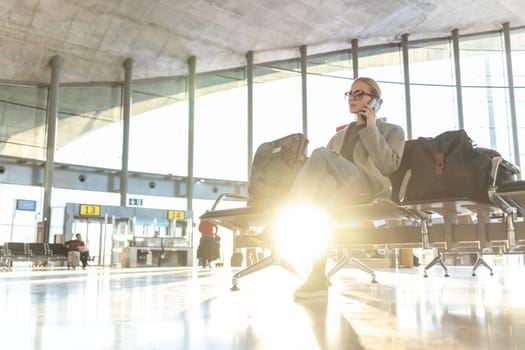 Fashionable blond young woman talking on her mobile phone while waiting to board a plane at the departure gates at the airport terminal.
