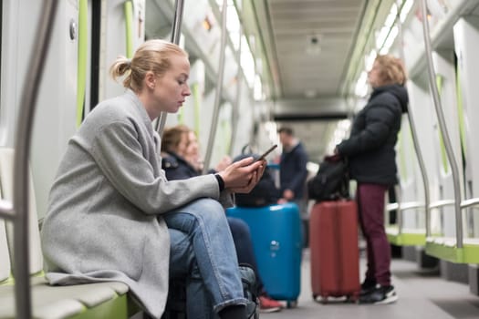 Beautiful blonde caucasian woman wearing winter coat reading on the phone while traveling by metro. Public transportation concept.