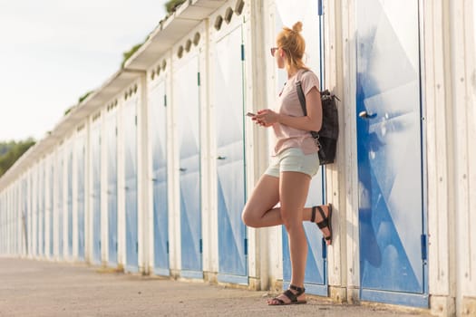 Blonde young female traveler wearing summer style clothing, holding mobile phone, against retro blue beach dressing rooms at summer time vacation in Sistiana, Italy.