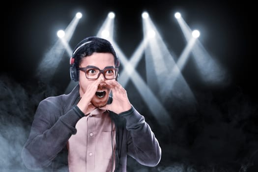 Expressive asian man in glasses wearing brown jacket enjoy the music with headphones over smoke and concert spot lighting background