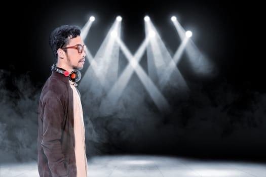 Young asian man in glasses wearing brown jacket with headphones on his neck standing over smoke and concert spot lighting background