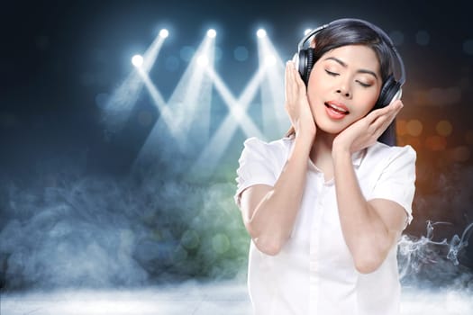 Smiling asian woman in white shirt listening music with headphones over concert spot lighting and smoke with defocused colorful lights background