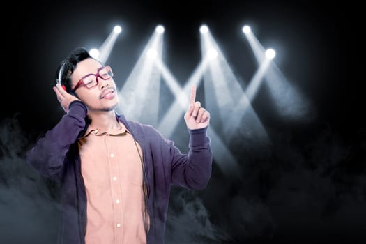 Happy asian man in glasses wearing brown jacket listen the music with headphones over smoke and concert spot lighting background