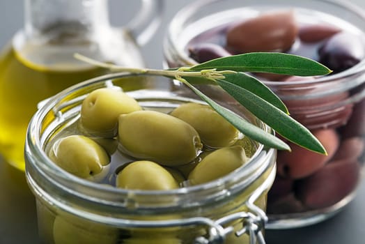Pickled green and black olives in glass jar with branch leaves close up