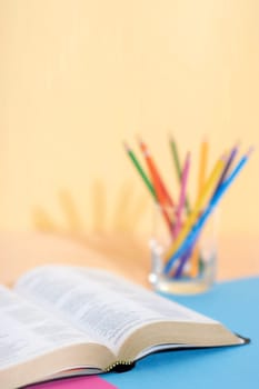 Bible book (blured text) and glass with a set of colored pencils on a colored paper background ready to be studied and underlined. Ready for Bible study.