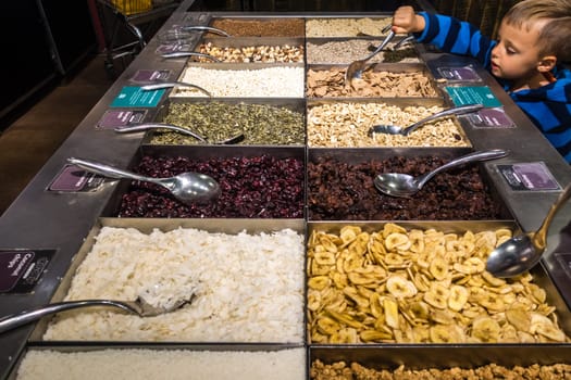 London, England - July 2018 : Young boy reaching out with a ladle to try the dehydrated fruits including raisins and pieces of coconut and banana on sale in the market stall