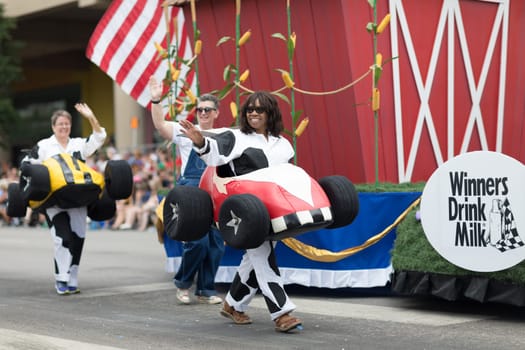 Indianapolis, Indiana, USA - May 26, 2018,  People with race car outfits walking along a float with a giant cow, promoting milk drinking, at the Indy 500 Parade