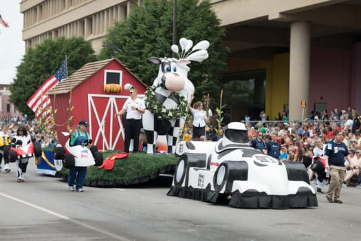 Indianapolis, Indiana, USA - May 26, 2018,  People with race car outfits walking along a float with a giant cow, promoting milk drinking, at the Indy 500 Parade