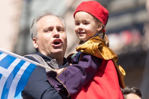 Chicago, Illinois, USA - April 29, 2018 Greek man hold his child wearing traditional clothing while the child waves the greek flag at the Greek Independence  Day Parade