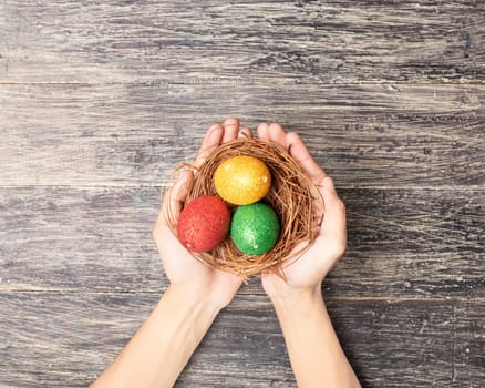 Human hands holding the nest with colorful easter eggs over wooden table background. Happy Easter