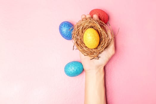 Human hand holding the nest with yellow easter egg and colorful easter eggs on pink color background. Happy Easter