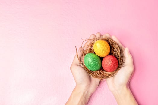 Human hands holding the nest with colorful easter eggs over pink color background. Happy Easter