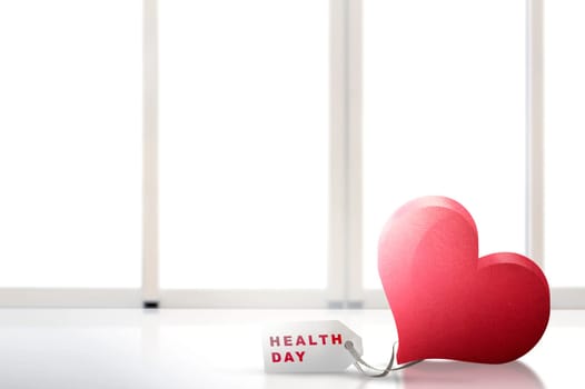 Red heart and a white paper tags on the table with Health day message with white window background. Health day concept