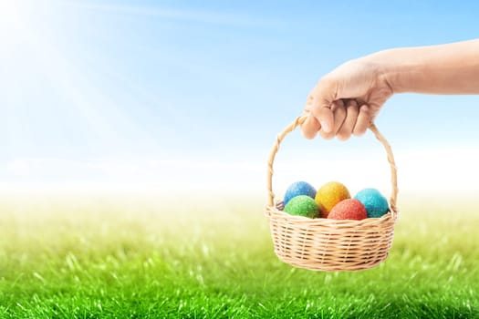 Human hands holding wooden basket with colorful easter eggs with green grass over blue sky background. Happy Easter