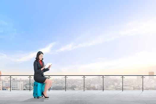 Beautiful asian business woman sitting on blue suitcase hold the tablet and work on modern terrace with city view and blue sky background