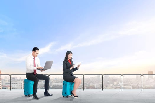 Young two asian business people sitting on blue suitcase hold the laptop and tablet working on modern terrace with city view and blue sky background