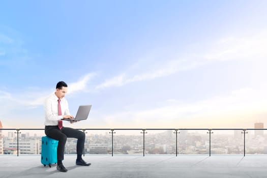 Handsome asian businessman sitting on blue suitcase hold the laptop and working on modern terrace with city view and blue sky background