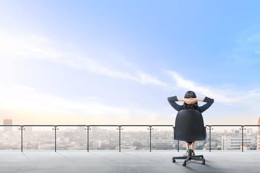 Rear view of asian business woman sitting on the office chair in modern terrace relax and looking at view on the city over blue sky background