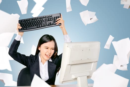 Stressed asian business woman with flying paper scattered want to slam the keyboard computer on the office room