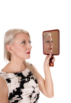 A middle age blond woman holding up a handheld mirror locking.at her makeup, standing in a dress, isolated for white background.
