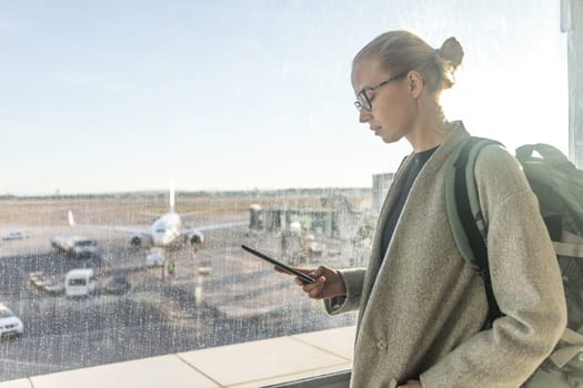 Casually dressed female traveler at airport looking at smart phone device in front of airport gate windows overlooking planes on airport runway.