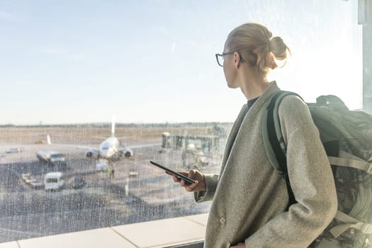 Casually dressed female traveler at airport, holding smart phone device, looking through the airport gate windows at planes on airport runway.